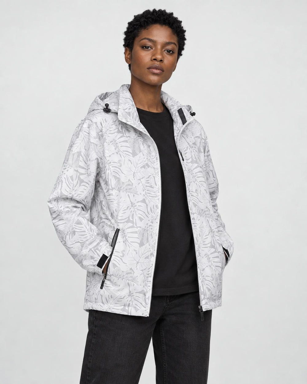 Model wearing the white leaf-print hooded windbreaker open over a black tee and black jeans, three-quarter view