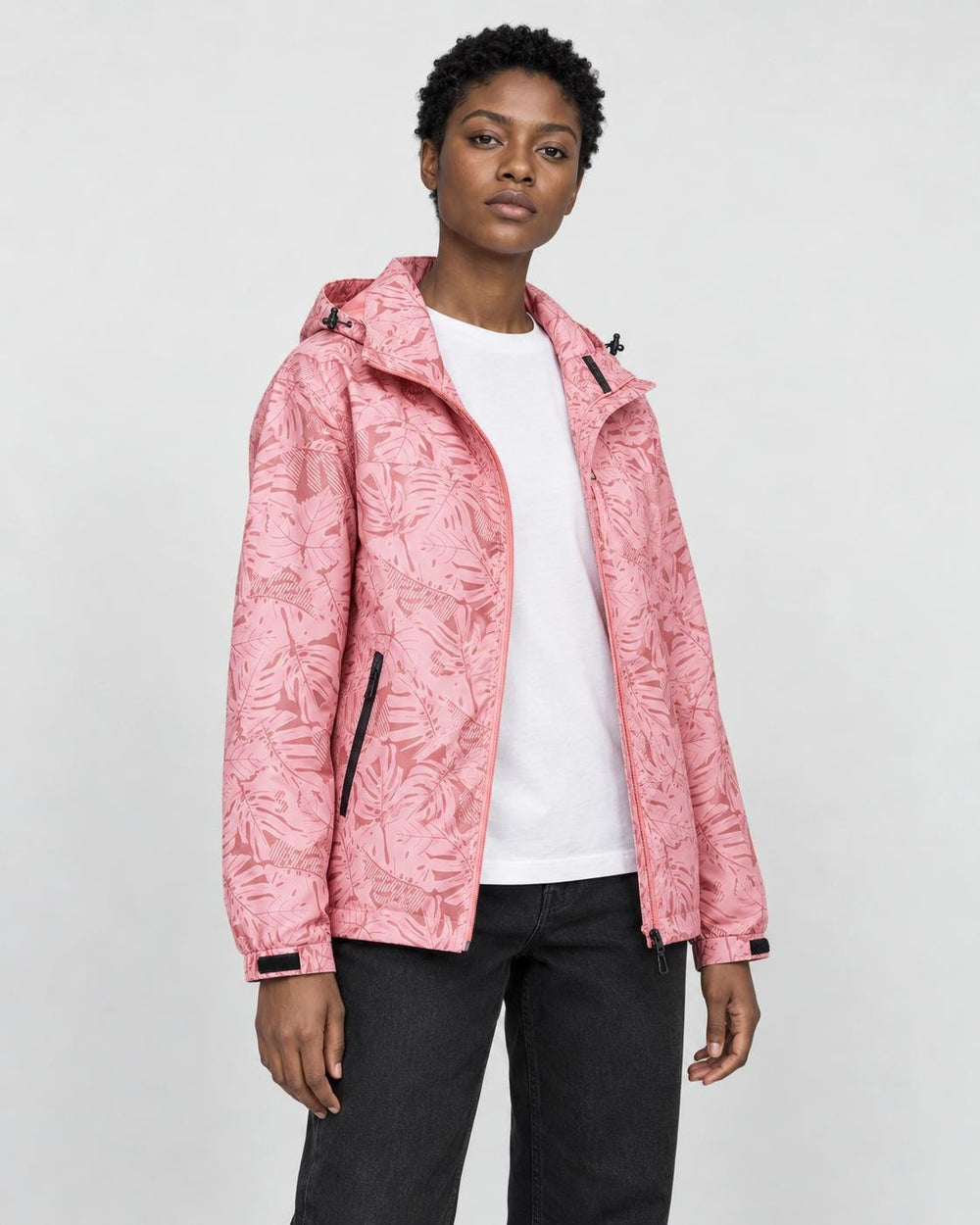 Model wearing the pink leaf-print hooded windbreaker open over a white tee and black jeans, three-quarter view