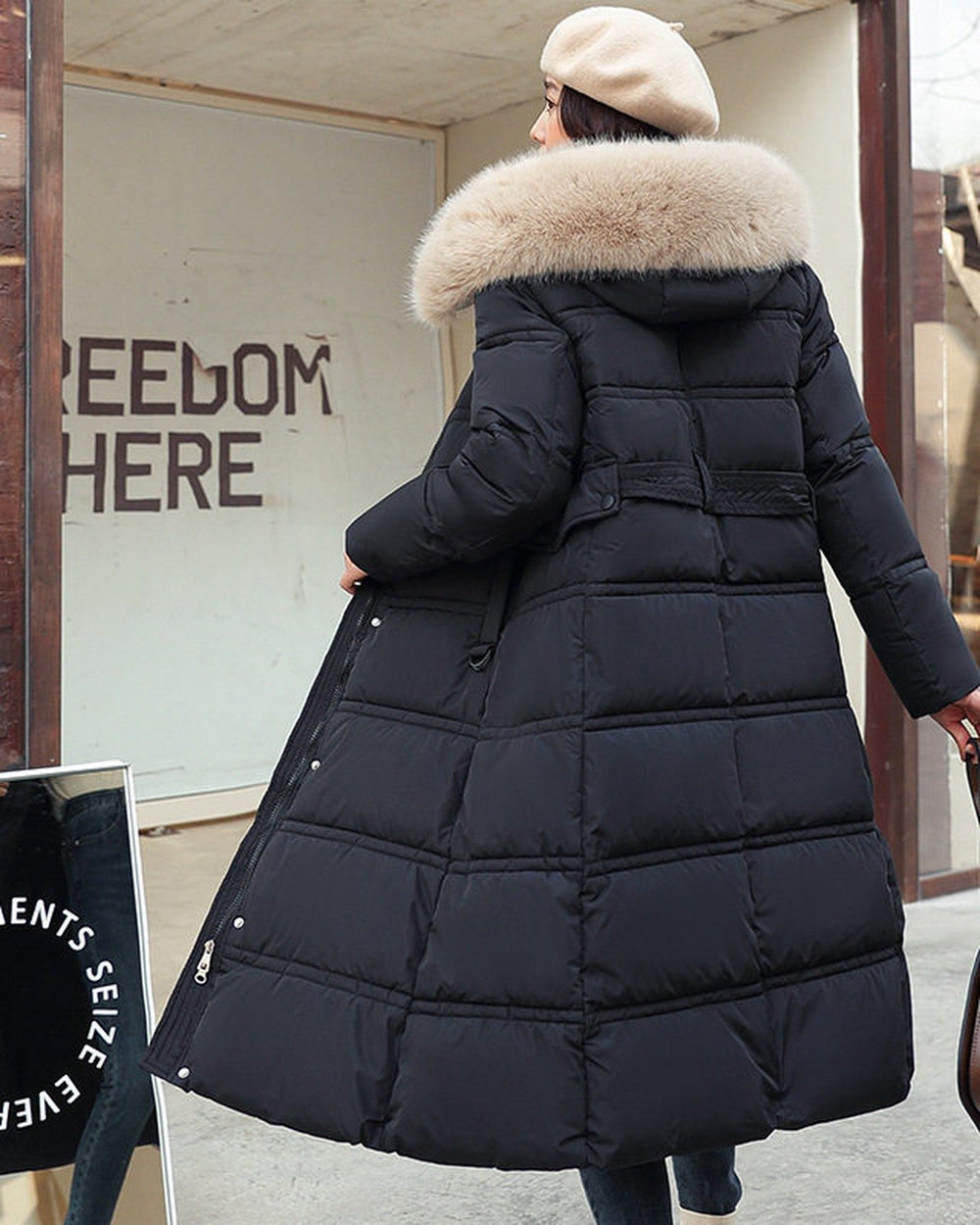 Black long puffer coat seen from behind with a khaki hood trim and a belt, worn with a brown shoulder bag