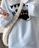 Light blue loose crew-neck sweatshirt with a black cat and lettering print, worn with a quilted bag