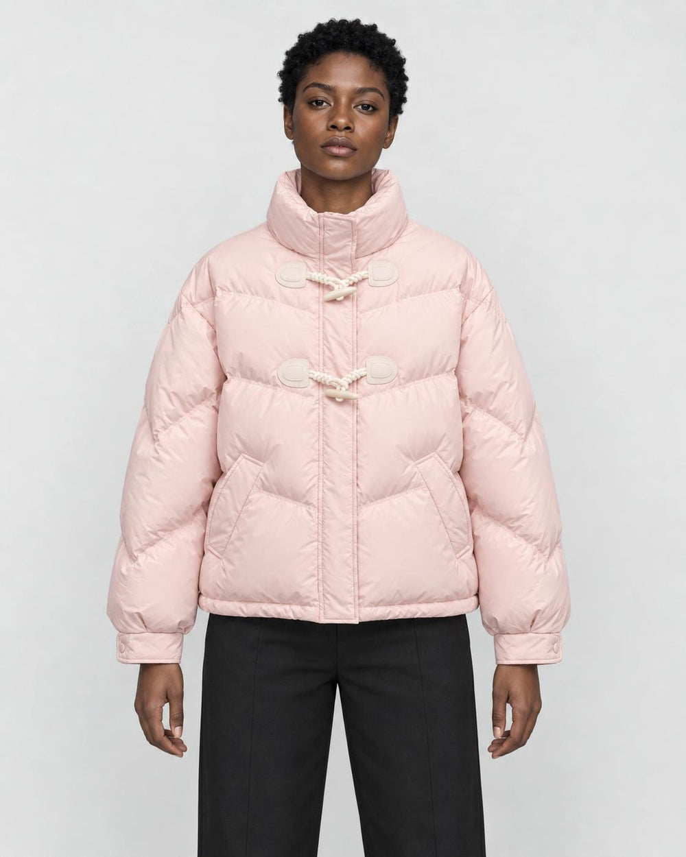 Woman in a short pink puffer jacket with horn toggles over a black top and black trousers, torso to mid-thigh